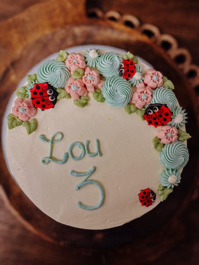Runder Kuchen mit Blumen- und Marienkäfer-Dekoration, beschriftet mit "Lou 3".