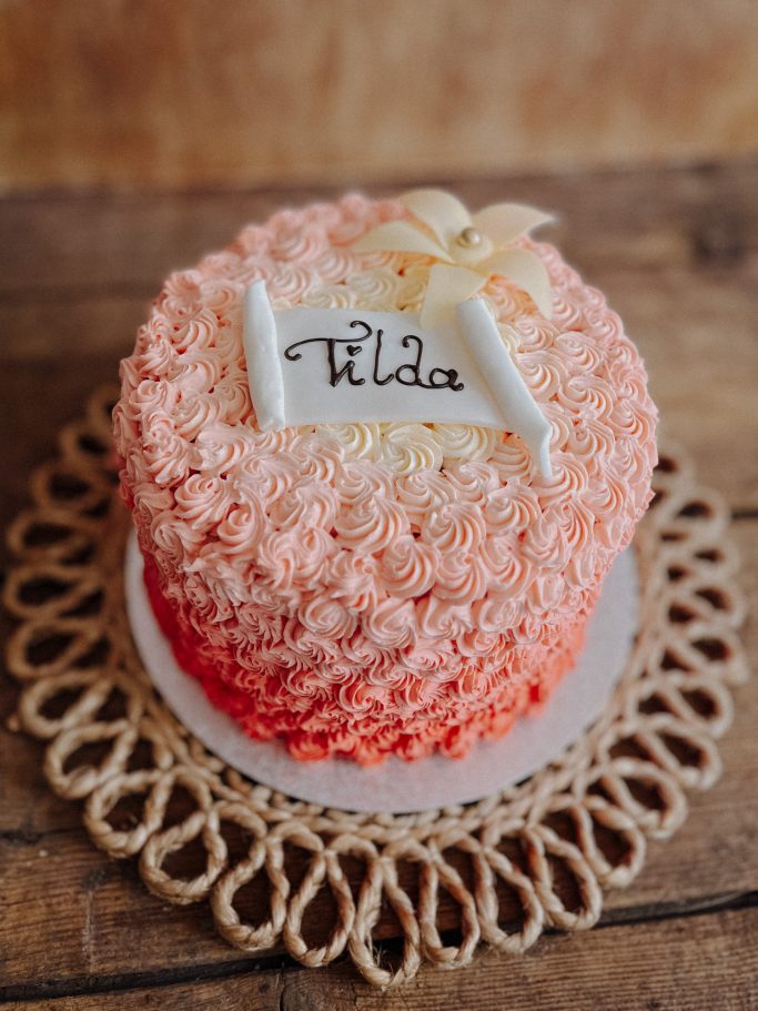 Rosa Ombre-Torte mit Blumenverzierung und "Tilde"-Schriftzug auf einem dekorativen Untersetzer.