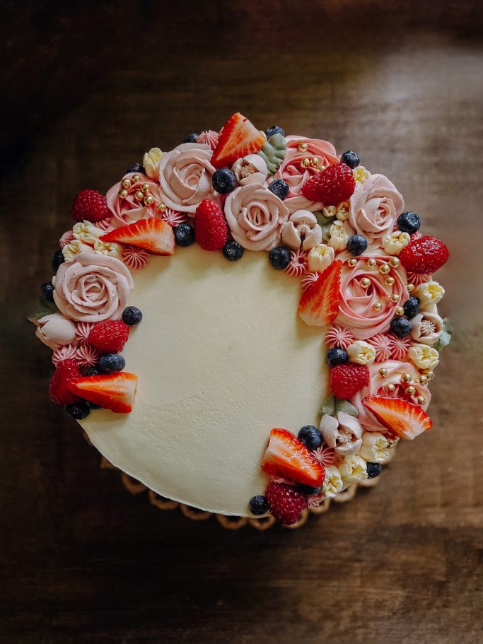 Kuchen dekoriert mit Rosen aus Sahne und frischen Beeren, auf einem Holzuntergrund.